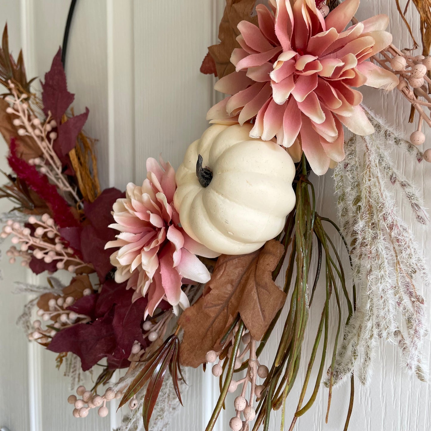 Close up of autumn hoop wreath with pampas grasses, maple leaves, pink berries, pink flowers and a neutral pumpkin. Styled on white door.