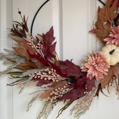 Close up of autumn hoop wreath with pampas grasses, maple leaves, pink berries, pink flowers and a neutral pumpkin. Styled on a white door.