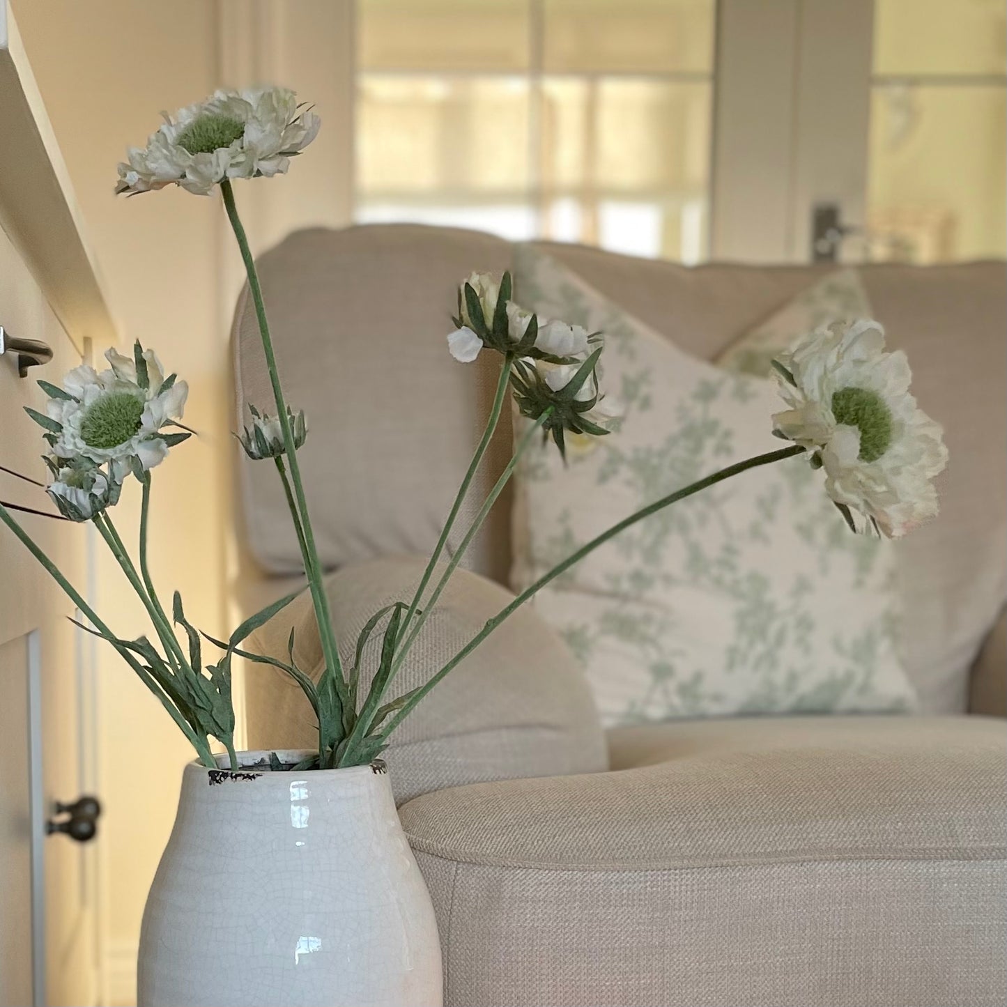 Close up of Faux Scabiosa Stem with off white flowers and soft green centres, styled in a Stephanie glazed vase. Styled in a cosy living room.