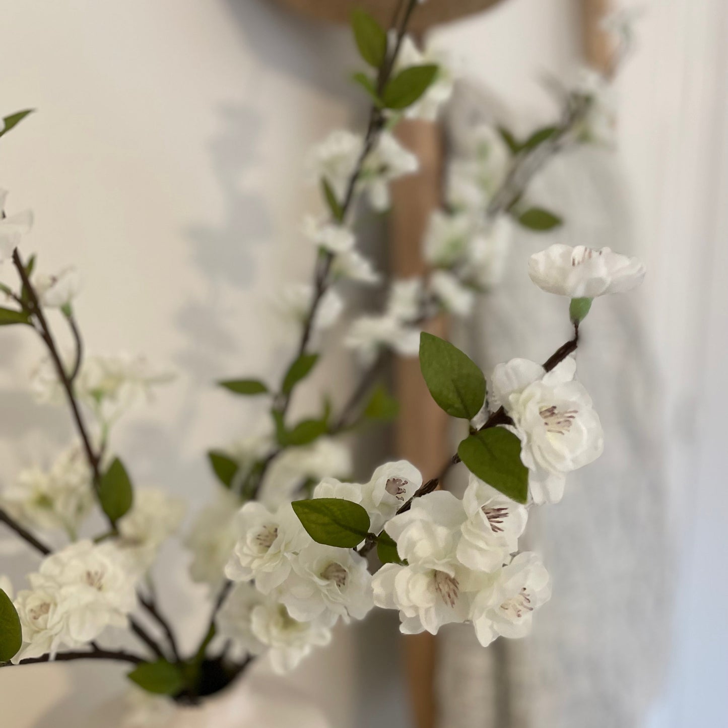 White faux blossom flowers with green leaves in a vase against a white background with a neutral throw.