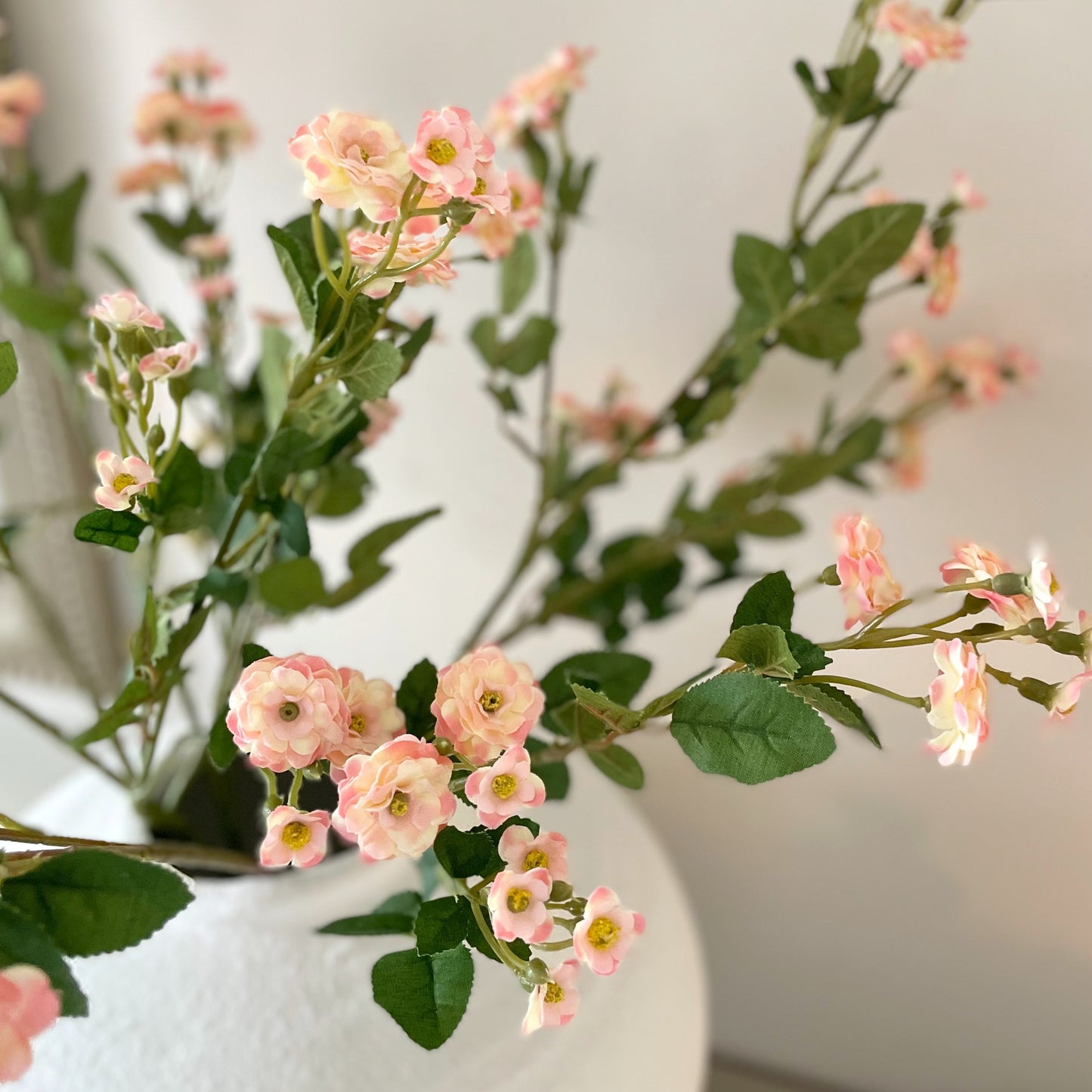 Wild Meadow Pink Rose Stem with delicate pink flowers, soft peach centres, and realistic green leaves. Styled in a white vase on a console table.