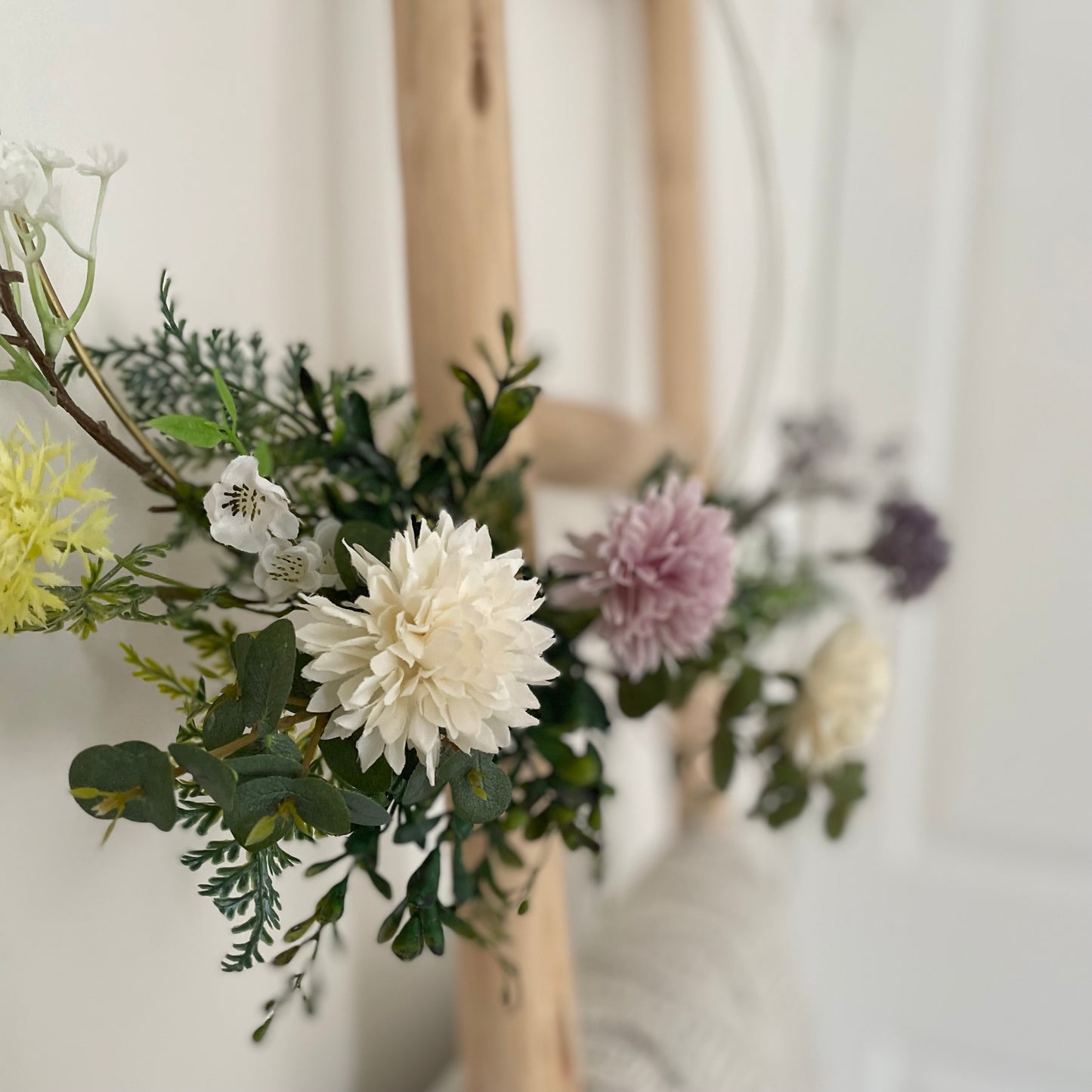 Whimsical hoop wreath decorated with artificial wild flowers and green foliage hanging on wooden ladder.