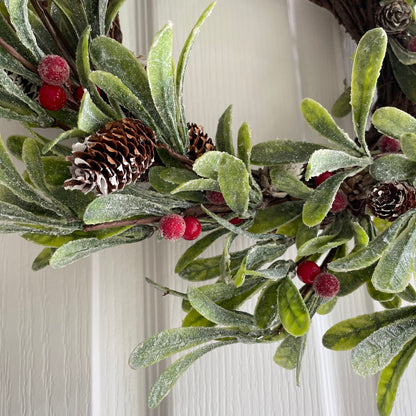 Frosted faux Christmas wreath with green foliage, red berries and fir cones, styled on a white wooden door.