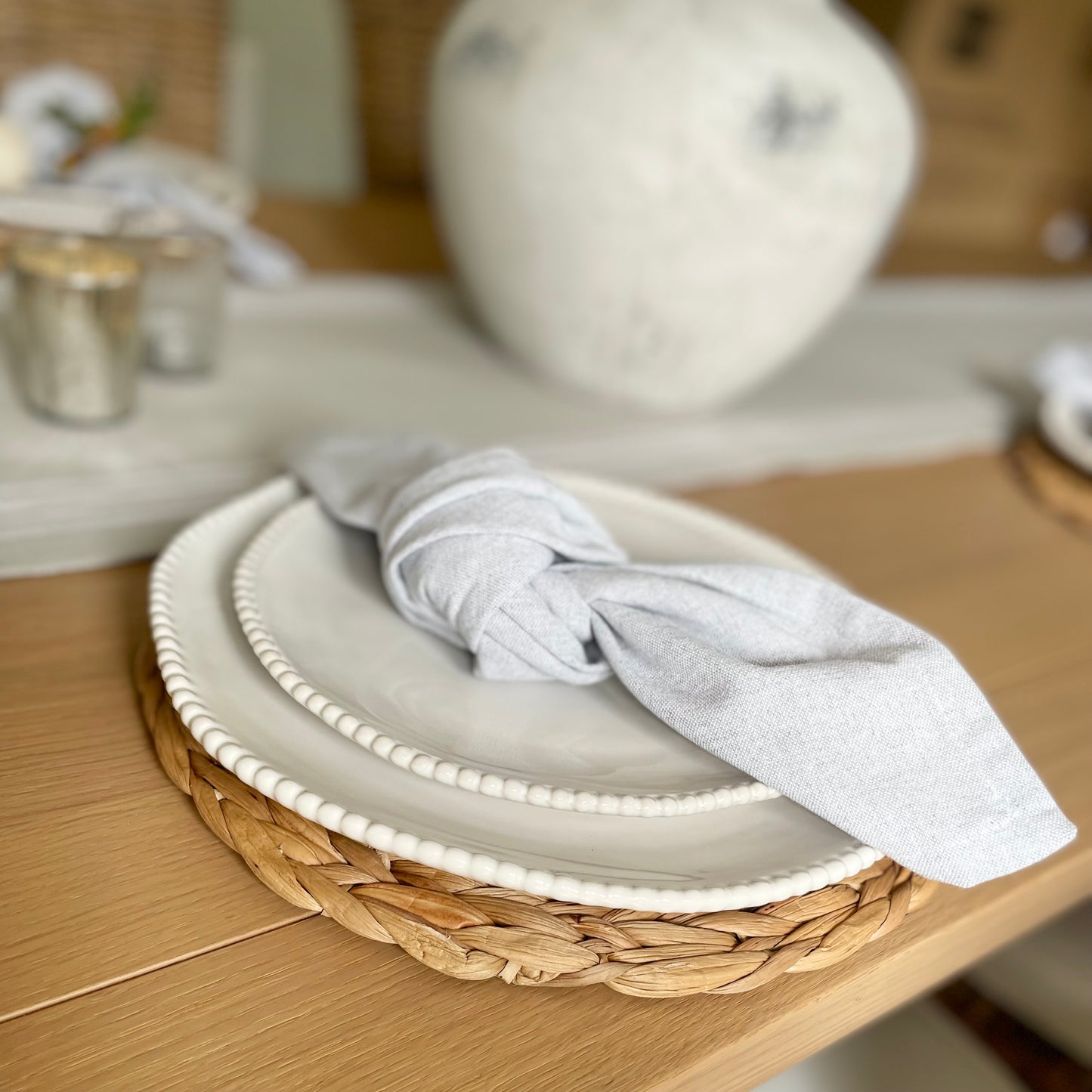 Set of four light grey cotton napkins neatly folded beside white porcelain plates and seagrass placemats on a dining table. 