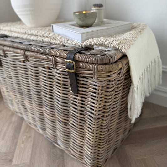 Wicker storage basket with a blanket draped over it on a wooden floor.