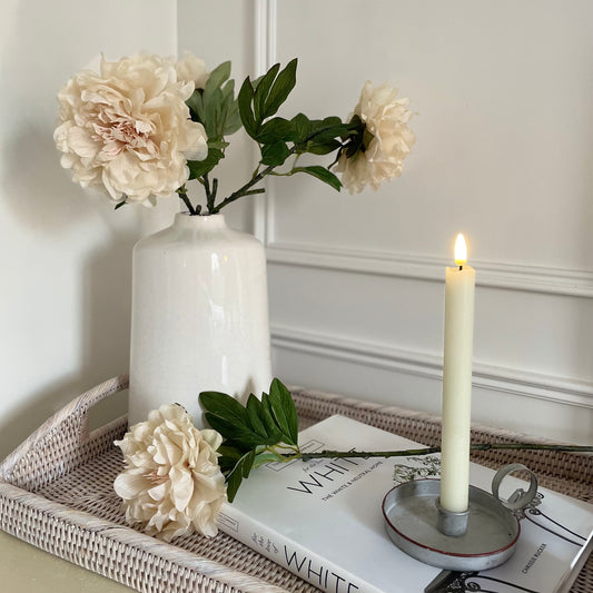 Soft faux peony stem with delicate cream petals and green leaves styled in a vase on a rattan tray with a candle.