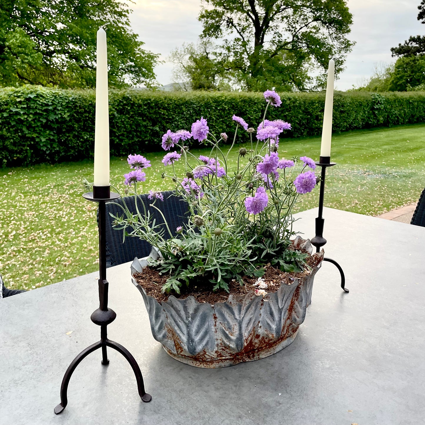 Tall rustic metal candlestick with thin stem and three leg base, styled on a dining table in the garden.
