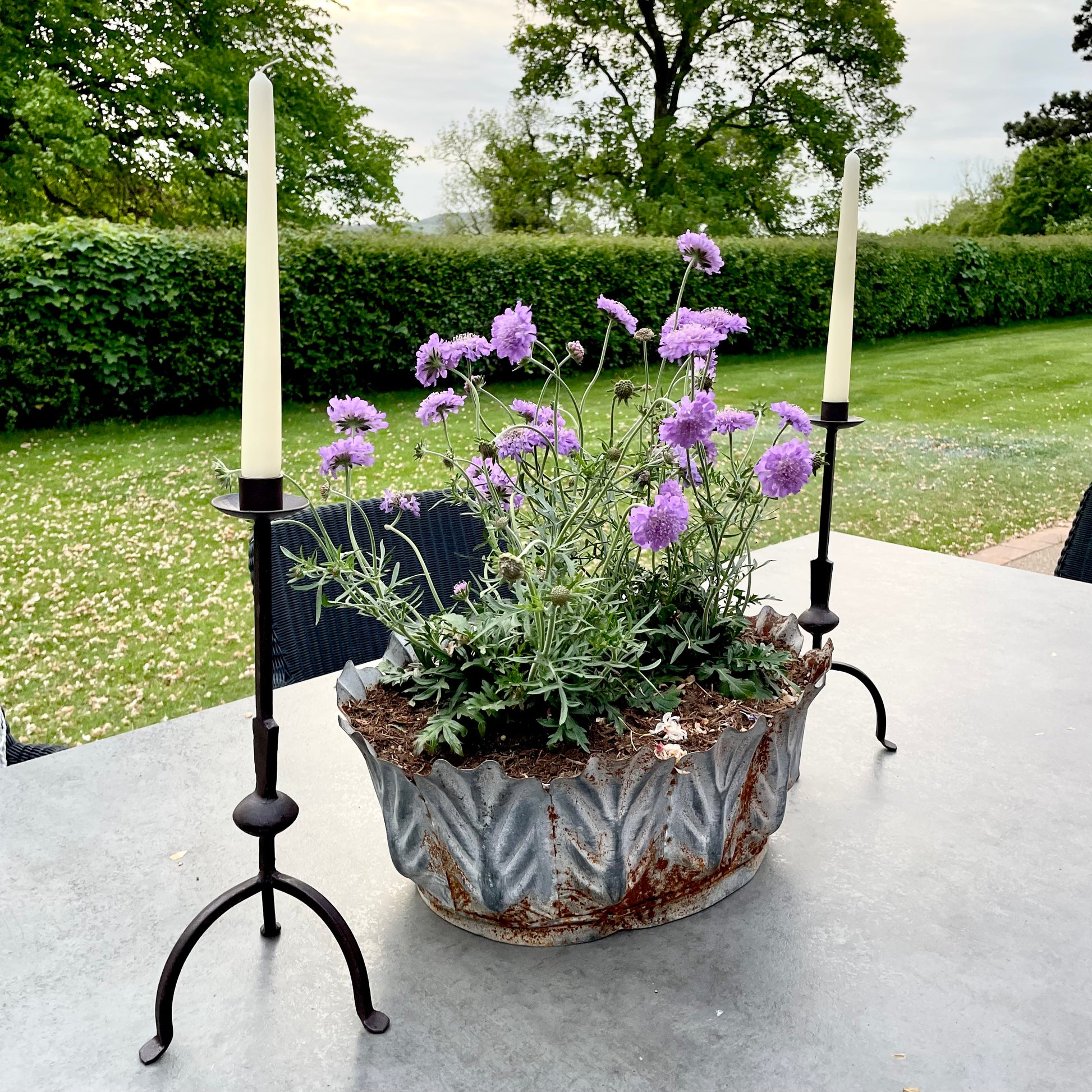 Tall rustic metal candlestick with thin stem and three leg base, styled on a dining table in the garden.