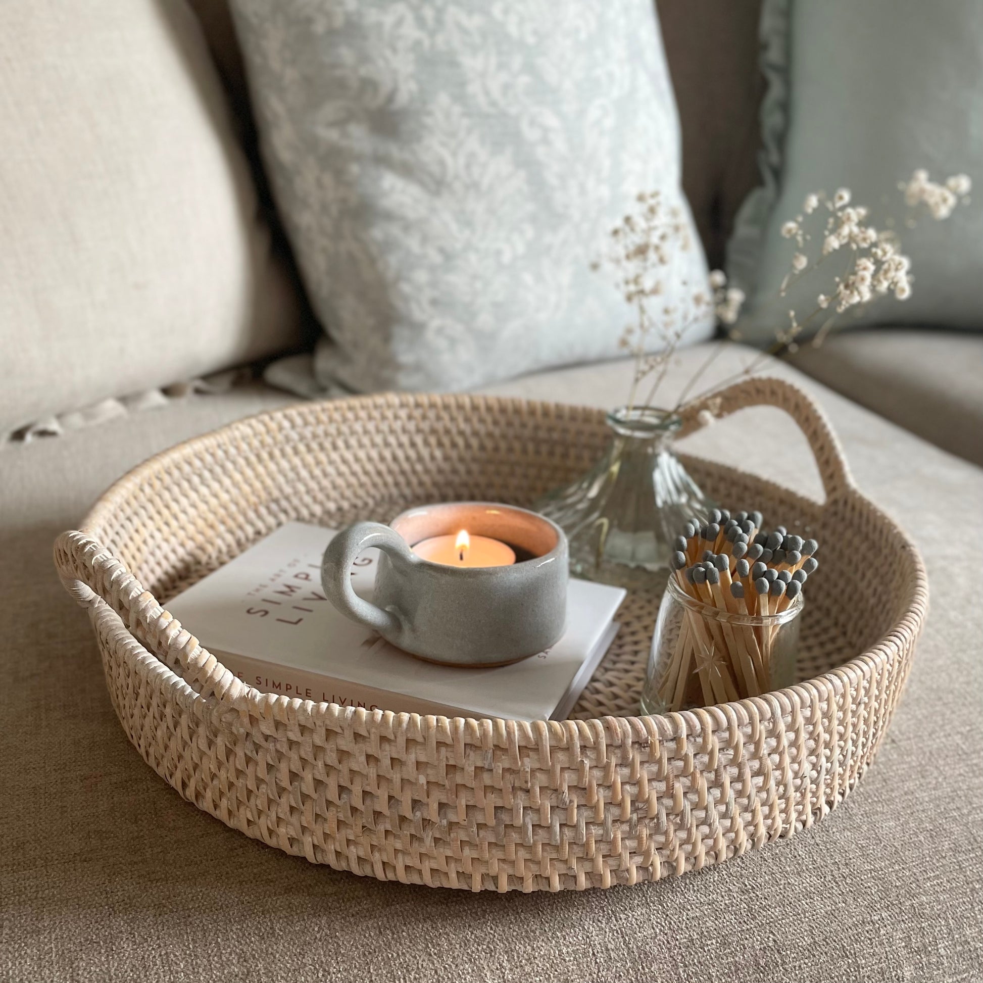 White ceramic tea light holder with handle, styled on a round rattan tray and book. In a cosy living room with plump cushions on a settee.