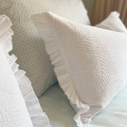 White quilted cushion with frilled edges and embroidered textured design. Styled on a bed.