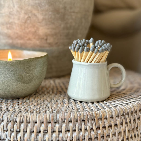 Matches in a small ceramic cup next to a lit candle on a woven surface