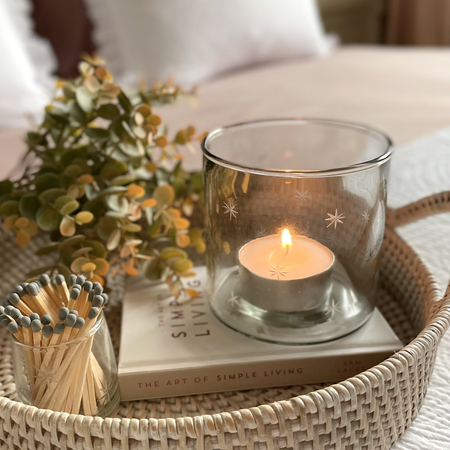 Candle in a etched star glass holder on a book with matches and greenery in a woven round basket.