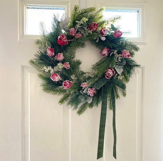 Handmade faux Christmas wreath with frosted red roses, flocked eucalyptus and a dusky green velvet bow on a natural grapevine base.