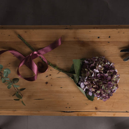 Faux hydrangea stem in shades of purple, burgundy and green styled on a rustic wooden table with burgundy ribbon.