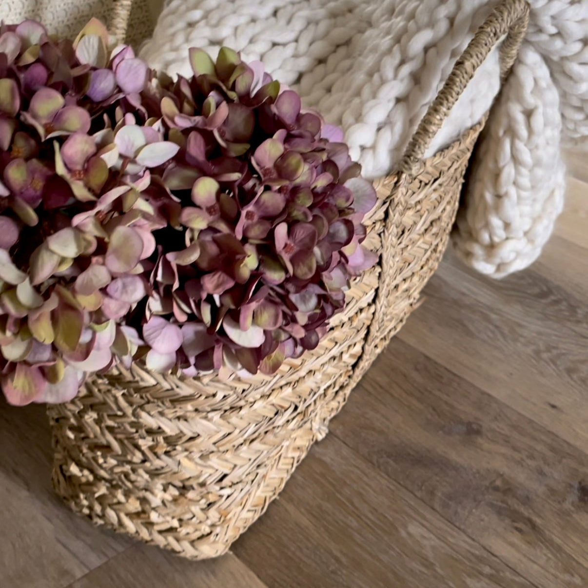 Close up of purple toned faux hydrangea stems in a woven seagrass basket, with a chunky cream knit throw. Styled on rustic wooden flooring.