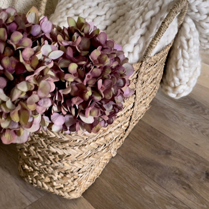 Close up of purple toned faux hydrangea stems in a woven seagrass basket, with a chunky cream knit throw. Styled on rustic wooden flooring.