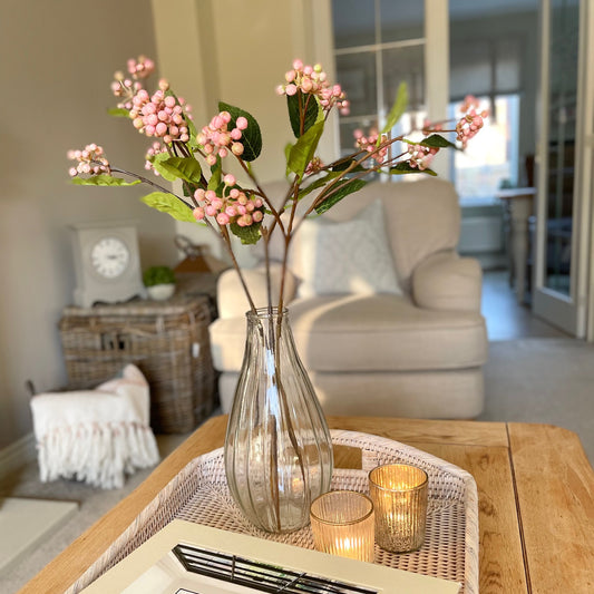 Faux pink berry stem with realistic clustered berries and green foliage, styled in a glass vase. With a rattan tray and candle on a coffee table in a cosy living room,