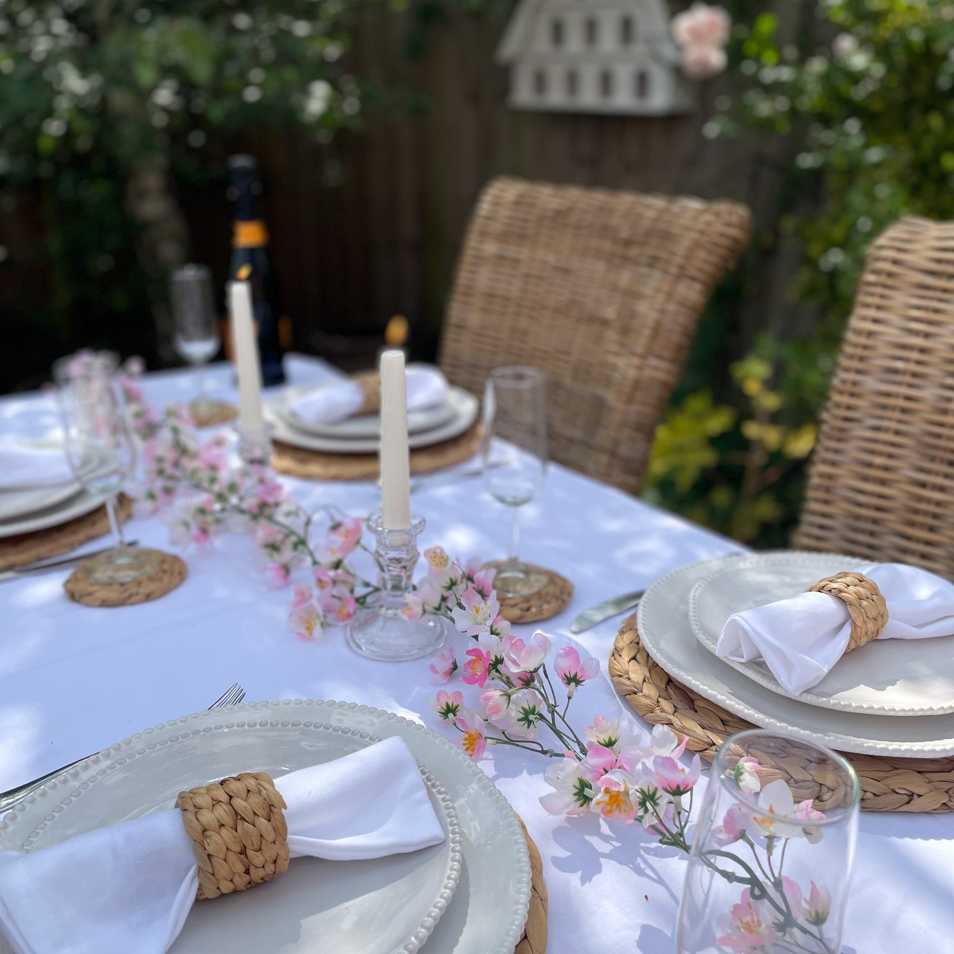 Pink cherry blossom garland with delicate lifelike petals and hanging loops. Styled on a garden table in a rose garden. Cosy inviting space with candles.