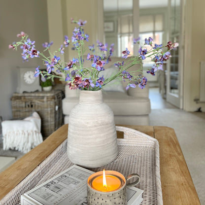 Faux blue and purple delphinium spray, 76 cm tall, styled in a stone vase on a rattan tray in a cosy living room.
