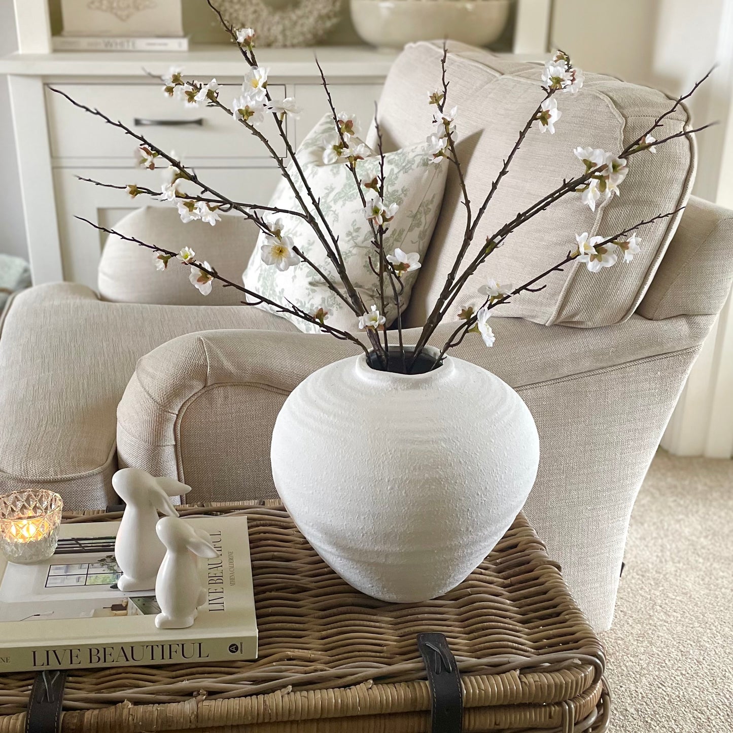 Artificial cherry blossom stem for floral arrangements. Styled in a white vase on a wicker trunk.