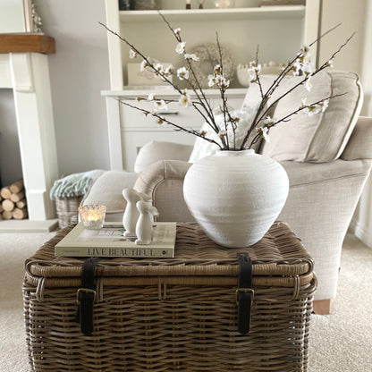 Faux cherry blossom branch with delicate white flowers and yellow pistils. Styled on a wicker trunk in a cosy living room.