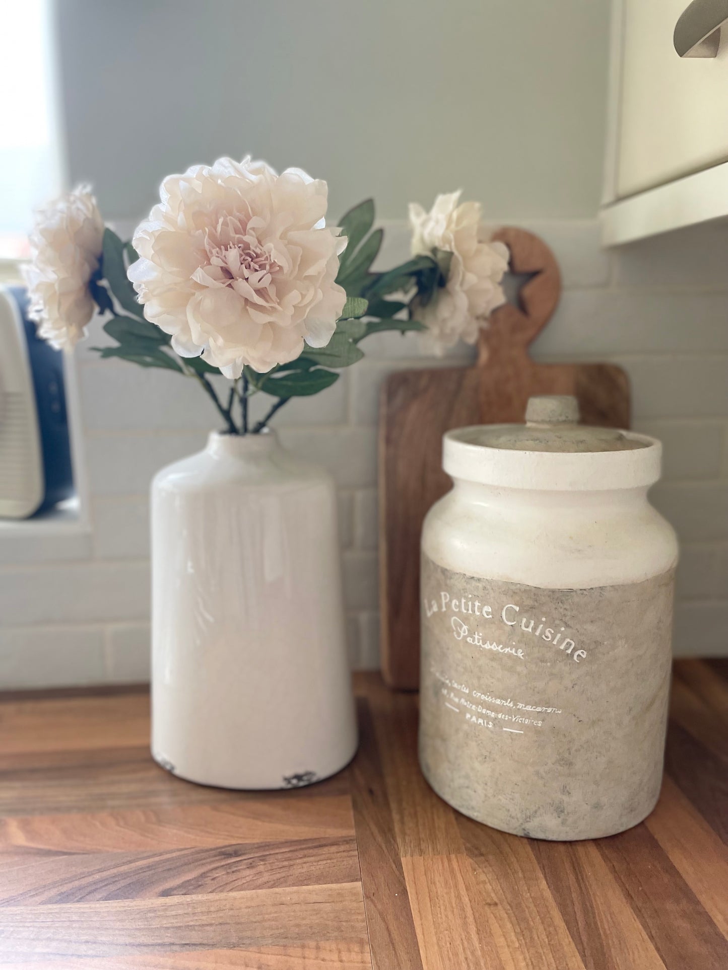 Soft faux peony stem with delicate cream petals and green leaves styled in a vase. Styled on a wooden kitchen worktop, with a ceramic storage pot.