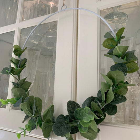 White metal wreath decorated with artificial eucalyptus and hanging on a door with jute string.