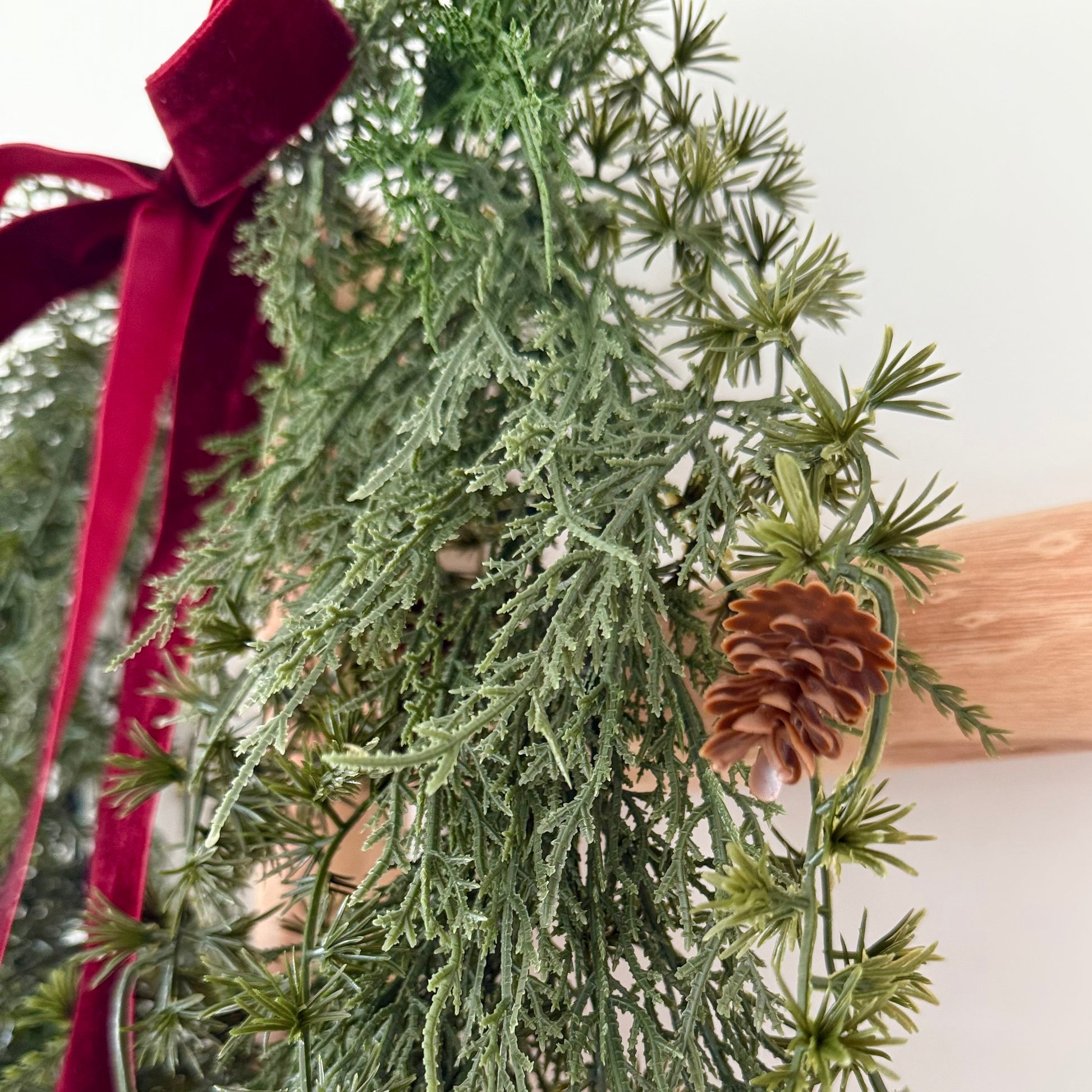 Faux green wreath with mini pine cones and a red ribbon on a white background.