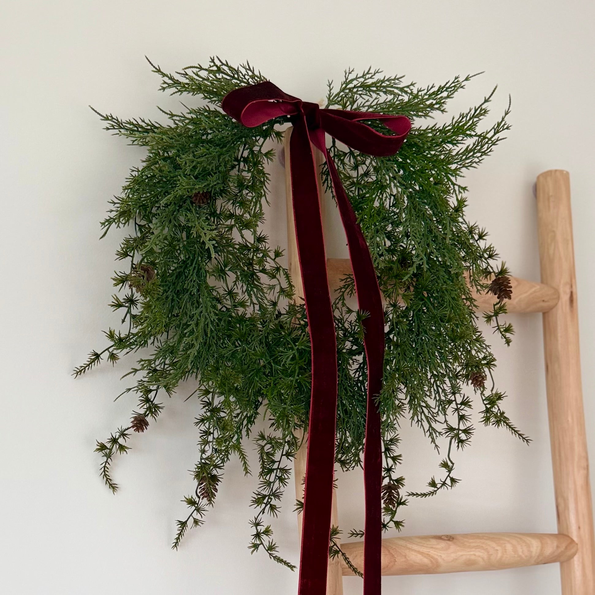 Green wreath with mini pinecones and a burgundy ribbon on a wooden ladder against a white wall