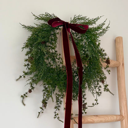 Green wreath with mini pinecones and a burgundy ribbon on a wooden ladder against a white wall