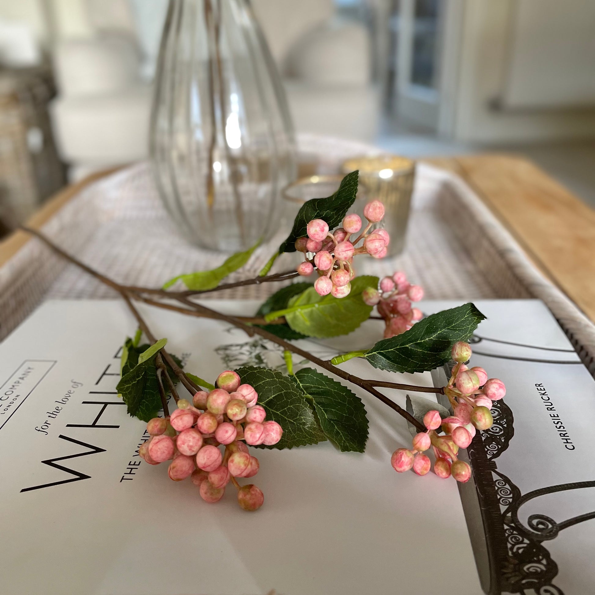 Faux pink berry stem with realistic clustered berries and green foliage, styled in a vase. On a rattan tray with a book.