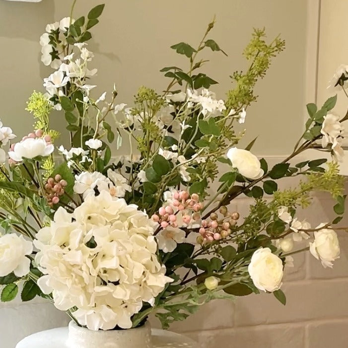 Faux spring bouquet with white ranunculus, wild roses, pink berries, mirabella sprays and a white hydrangea. Styled in a vase on a wooden kitchen worktop with whie tiled background.