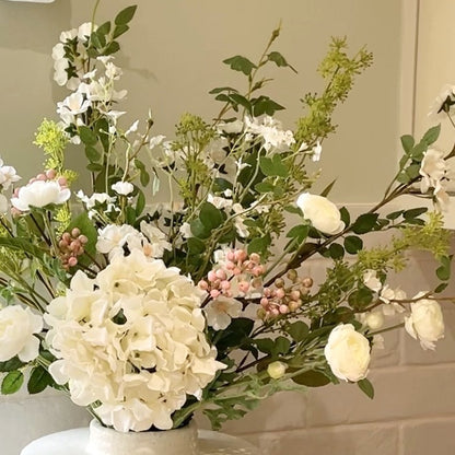 Faux spring bouquet with white ranunculus, wild roses, pink berries, mirabella sprays and a white hydrangea. Styled in a vase on a wooden kitchen worktop with whie tiled background.