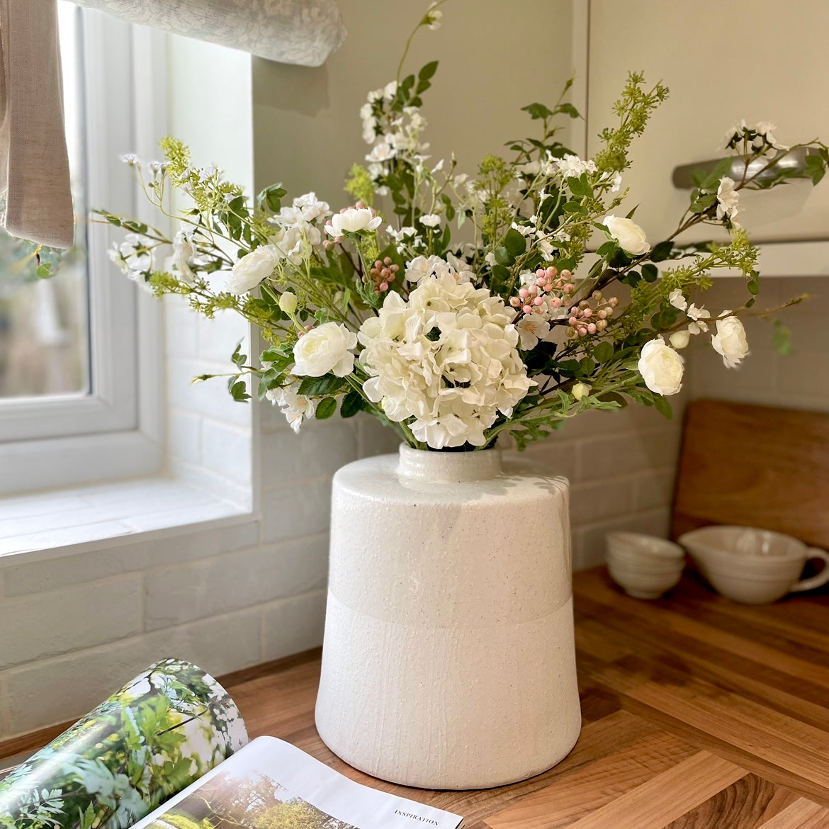 Faux spring bouquet with white ranunculus, wild roses, pink berries, mirabella sprays and a white hydrangea. Styled in a vase on a wooden kitchen worktop.