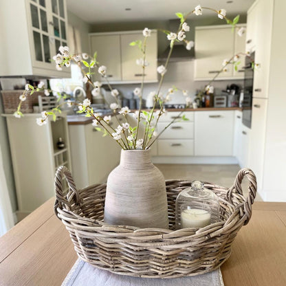 Faux white blossom spray with three stems styled in a stone vase in a rattan basket on a dining table.