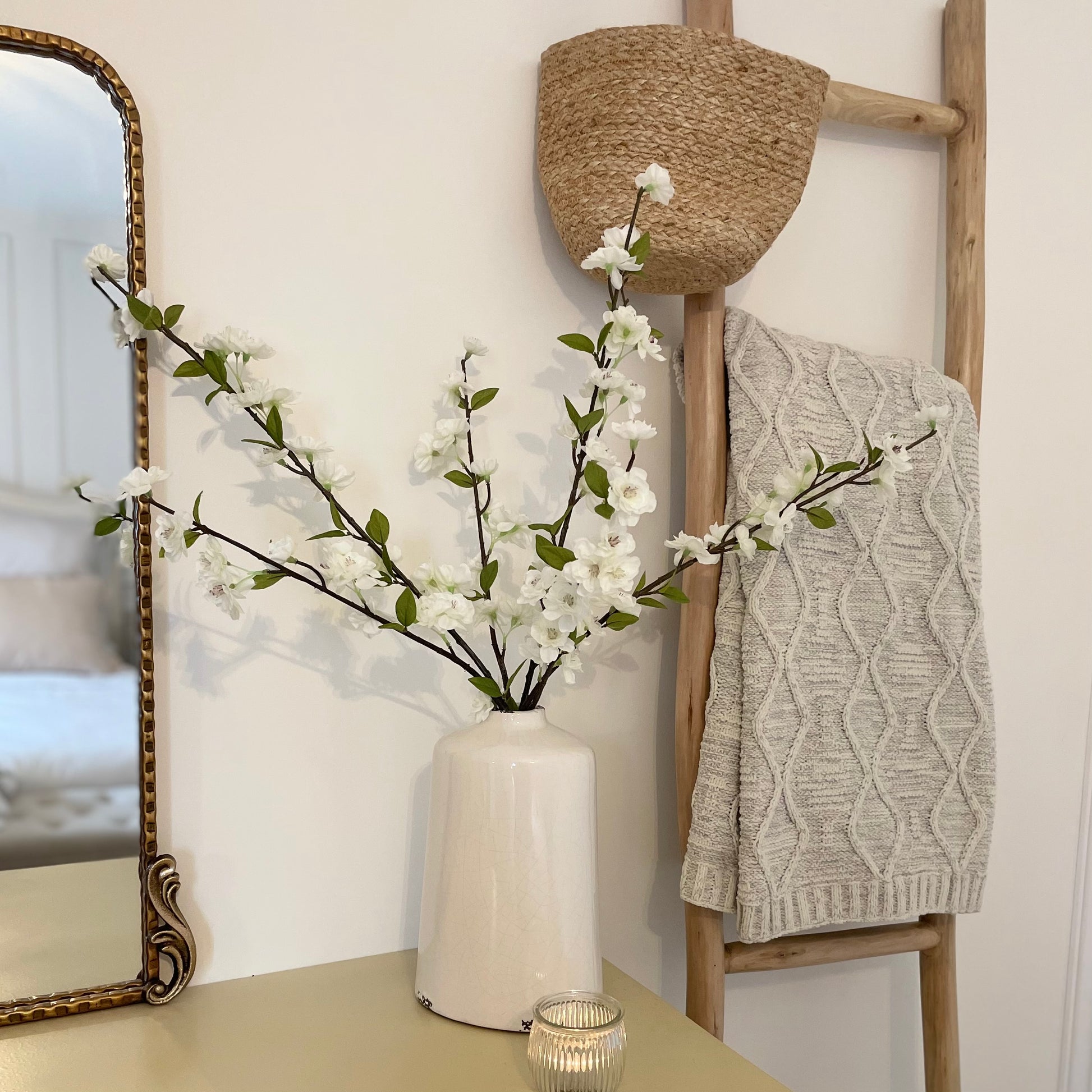 Delicate faux white blossom stems with green leaves displayed in a ceramic vase on a dressing table with a candle.
