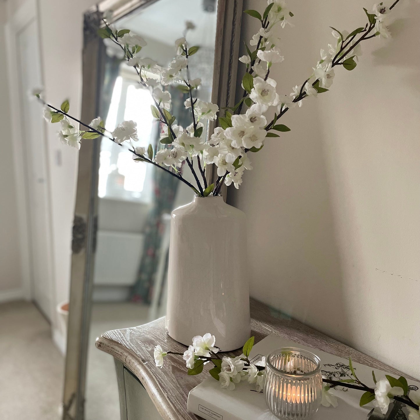 Ceramic vase with faux white blossom flowers, styled on a console table in a hallway with a lit candle. 