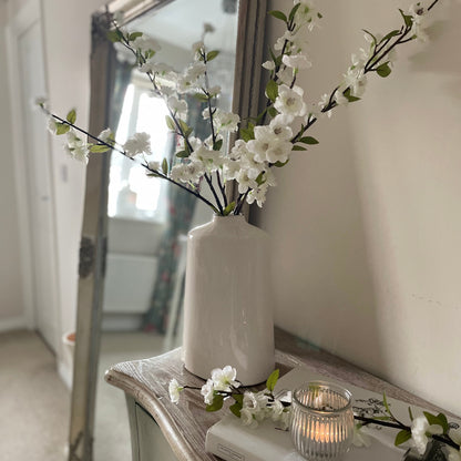 Ceramic vase with faux white blossom flowers, styled on a console table in a hallway with a lit candle. 