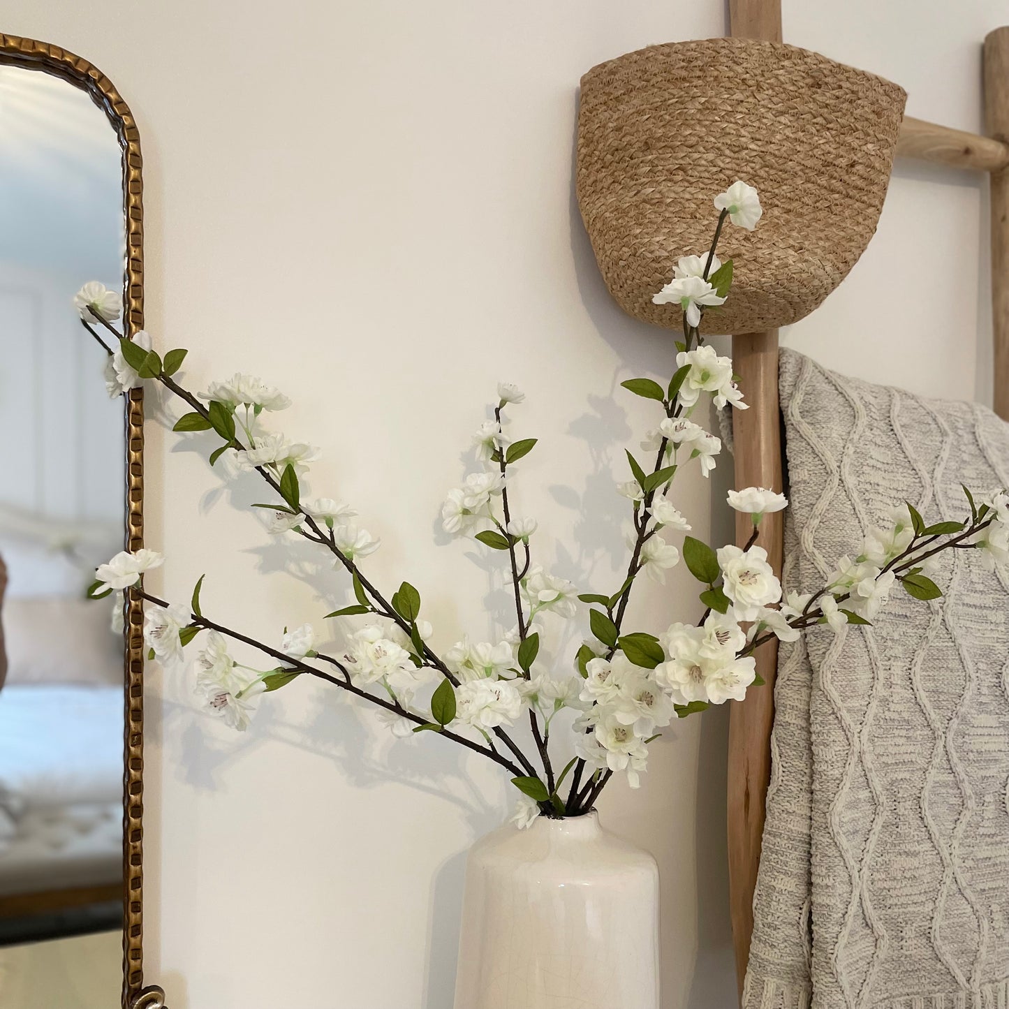 Delicate faux white blossom stems with green leaves displayed in a ceramic vase. With a cable knit throw and hanging basket.