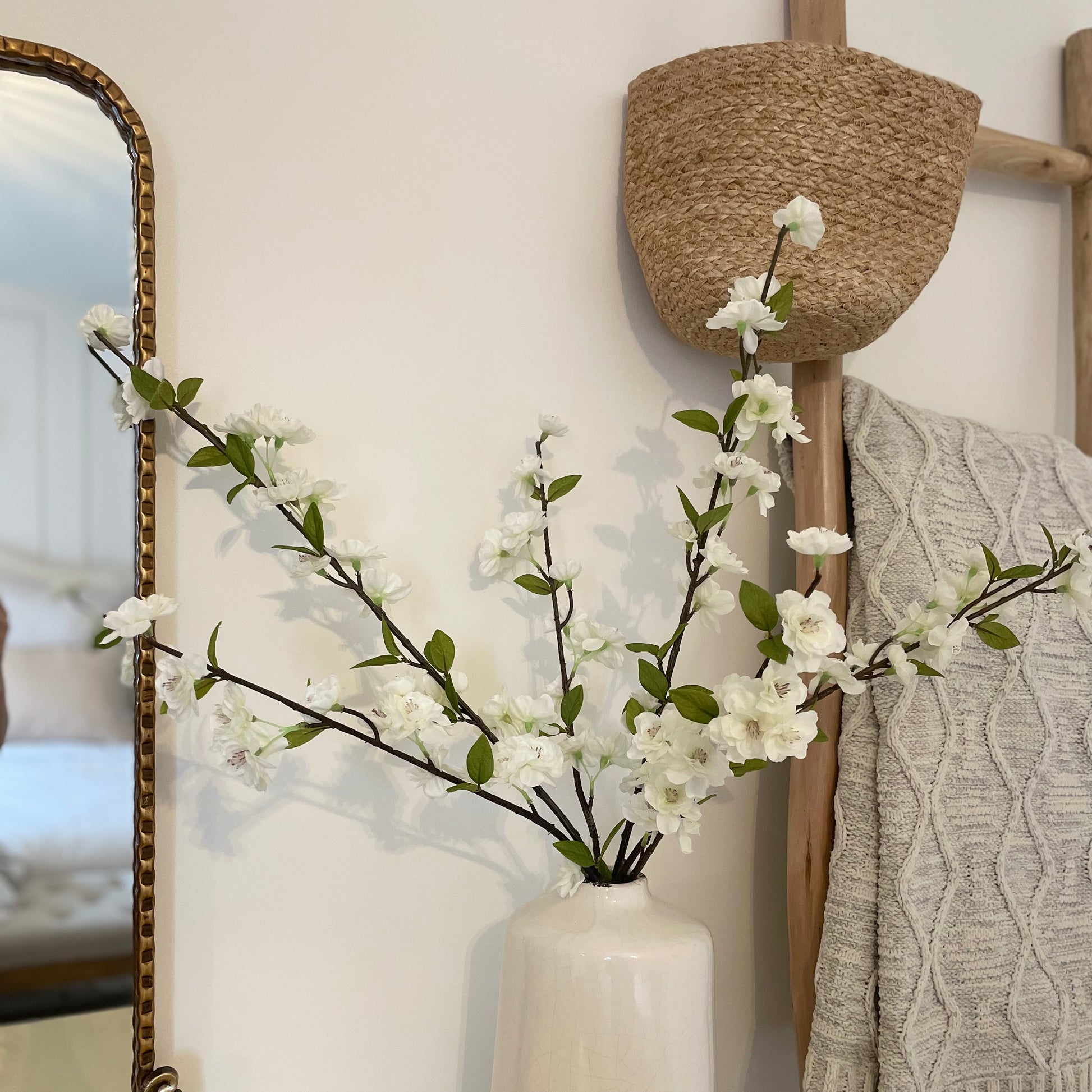 Delicate faux white blossom stems with green leaves displayed in a ceramic vase. With a cable knit throw and hanging basket.