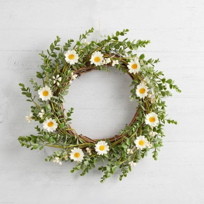 Faux daisy wreath with white flowers and green foliage. Styled on a white wooden wall.