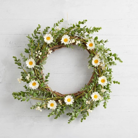 Faux daisy wreath with white flowers and green foliage. Styled on a white wooden wall.