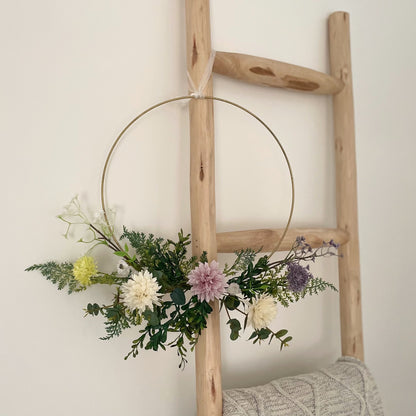 Whimsical hoop wreath decorated with artificial wild flowers and green foliage hanging on wooden ladder with a cable knit throw.