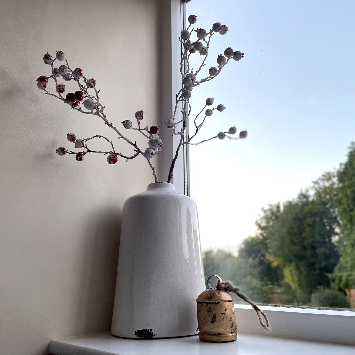 Faux winter red berry stem styled in a ceramic white vase. Displayed on a windowsill with a brass decorative bell.