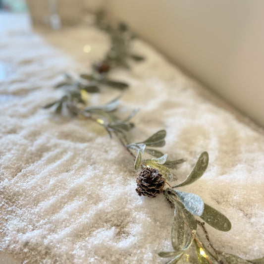 Frosted pinecone garland with miniature cones and glitter. Displayed on a snowy background.