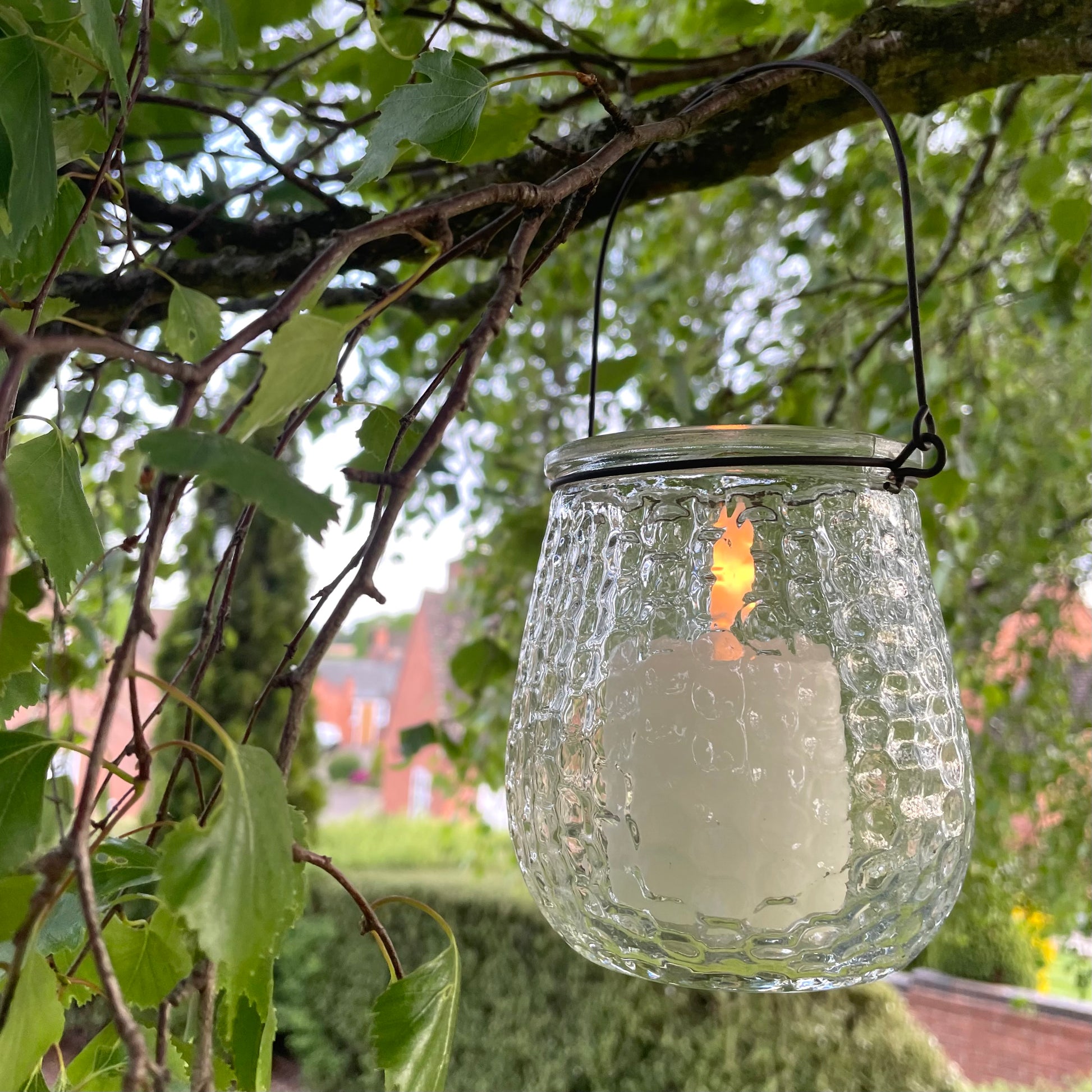 Clear glass hanging candle holder with metal handle, styled outdoors with a glowing tea light.
