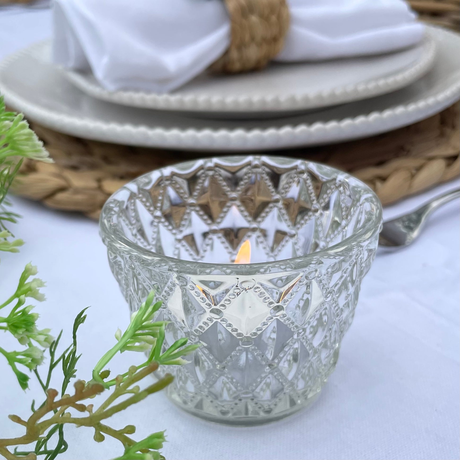 Clear glass tea light holder with an embossed diamond beaded pattern, styled on a table with soft, glowing candlelight.