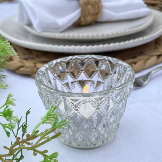 Clear glass tea light holder with an embossed diamond beaded pattern, styled on a table with soft, glowing candlelight.