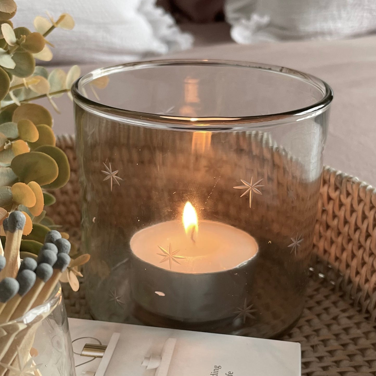 Glass hurricane candle holder with delicate etched star design, styled with a glowing candle on a rattan round tray.