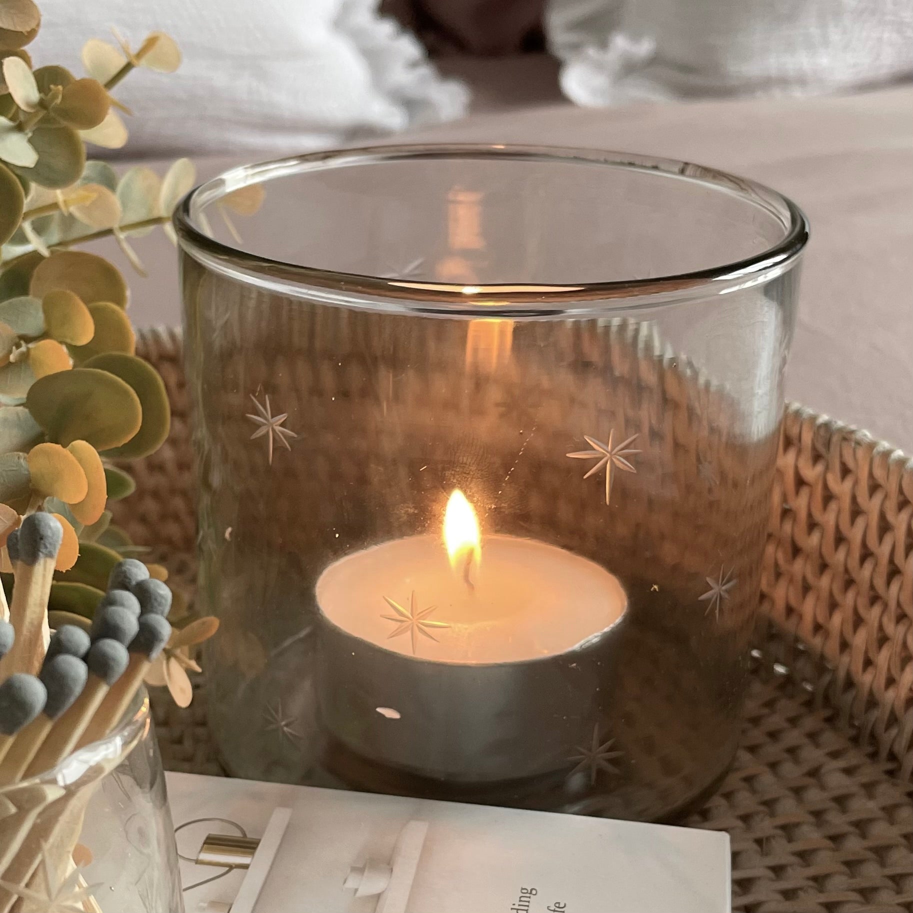 Glass hurricane candle holder with delicate etched star design, styled with a glowing candle on a rattan round tray.